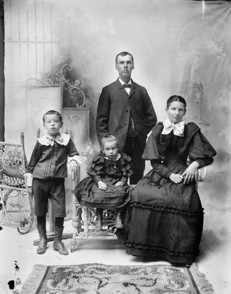 Studio portrait in front of a painted backdrop of a man, woman, and two children. The man in the center is wearing a watch fob on his vest and is standing behind the group. On the left is a young boy wearing high button boots and a white collar and cuffs, and who is standing and leaning his left arm on the arm of a wicker bench. On the bench is a young girl wearing a dress, high button boots and a locket, and sitting in the center. On the right side of the bench is a woman sitting who is wearing a full-length dark dress with ruffles and puffy sleeves, and a large white bow at her neck.
