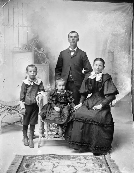 Studio portrait in front of a painted backdrop of a man, woman, and two children. The man in the center is wearing a watch fob on his vest and is standing behind the group. On the left is a young boy wearing high button boots and a white collar and cuffs, and who is standing and leaning his left arm on the arm of a wicker bench. On the bench is a young girl wearing a dress, high button boots and a locket, and sitting in the center. On the right side of the bench is a woman sitting who is wearing a full-length dark dress with ruffles and puffy sleeves, and a large white bow at her neck.