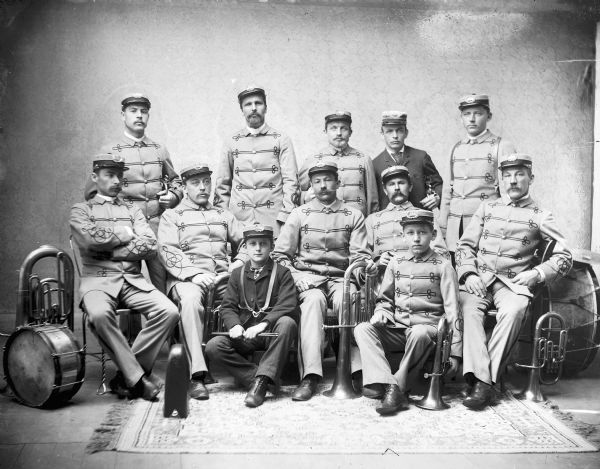 Studio portrait of band members, posing in their uniforms and with their instruments. Two young boys are sitting in the front of the group. The insignia on the caps they are all wearing reads: "ABB."