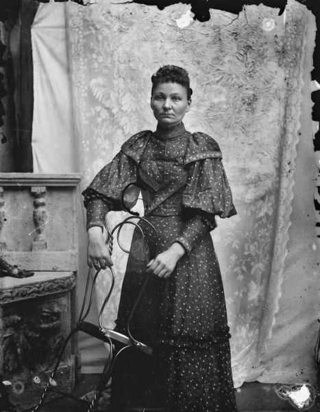 Studio portrait of an unidentified woman wearing a full-length dark-colored dress with polka-dots, embellished with leaf patterned trim, ruffles and puffy sleeves. She is holding a wrought iron chair tilted towards her, and in the background is a lace curtain over a white backdrop.
