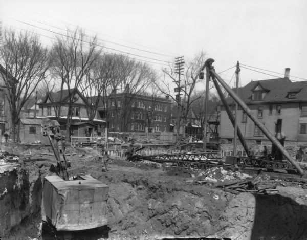 Orpheum Theater construction, 216 State Street, looking north, with steam shovel and other equipment. Also shows Holy Redeemer Catholic Church, 128 W. Johnson Street and Holy Redeemer Rectory, 120 W. Johnson Street, and Windemere Apartments, 118 W. Johnson Street.