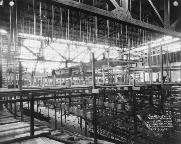 Interior view of Orpheum Theater construction, 216 State Street, from upper balcony. A man is standing on top of scaffolding in the center. Below him coats are hanging from the scaffolding. Another man (blurred from movement) is working on the balcony on the far right. Caption reads: "Orpheum Theatre, Madison, Wisconsin, Beecroft Bldg. Co., Builders; C.W. & Geo. L. Rapp, Architects; A.D. & J.V. Fredrickson, Contractors."