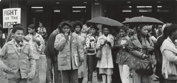 Several African-American teenagers are standing outside a building, some wearing coats and some under umbrellas. A sign reads: "Fight for the Right." Caption reads: "Some of the estimated 1,300 youngsters who participated Friday in a black pupil walkout from Milwaukee Public Schools left Parkman Junior High School. The walkout, which was sponsored by the Organization of Organizations and Blacks for Two Way Integration, was to protest desegregation and its burden on blacks."