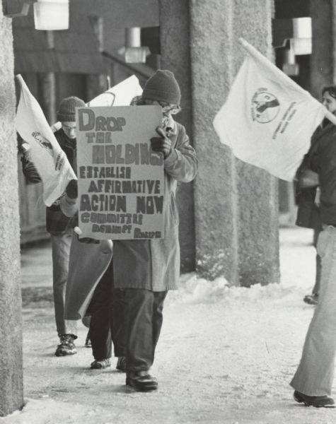 Several people wearing coats, hats, and gloves are walking in a circle outside a building. One person is holding a sign that reads: "Drop the Holdings, Establish Affirmative Action Now, Committee Against Racism." Other people are holding flags with the Committee Against Racism symbol.