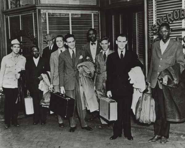 Nine men are posing in front of a storefront for S.W. Robinson Real Estate. They are holding suitcases and coats and are all wearing suits. Caption reads: "Richmond VA 1947. Group of participants in the Journey of Reconciliation sponsored in 1947 by the Fellowship of Reconciliation and the Congress of Racial Equality. Bayard Rustin is in the center rear, and George Houser is second from right in front, then executive secretary of CORE."