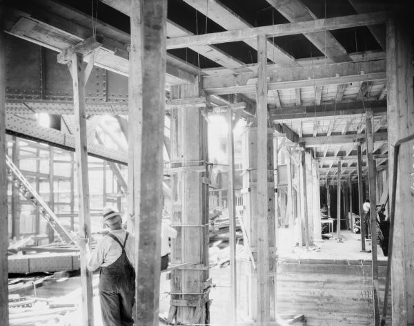Orpheum Theater construction, 216 State Street, looking southeast. A man is standing in the foreground with his back towards the camera, and another man (obscured by a beam) is sitting on a board nearby. A man is working on the far right in the background.