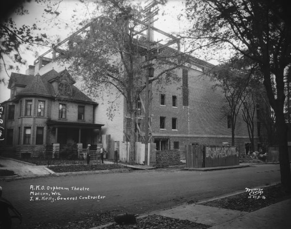 View across W. Johnson Street towards the back side of the Orpheum Theater under construction at 216 State Street. The house beside the theater on the left is at 117 W. Johnson Street. Men are standing in front of the house. A sign on a wooden fence along the theater reads: "Are you ready to take a chance at the Orpheum this week?" Text on print reads: "R.K.O. Orpheum Theatre, Madison, Wis., J.H. Kelly, General Contractor."