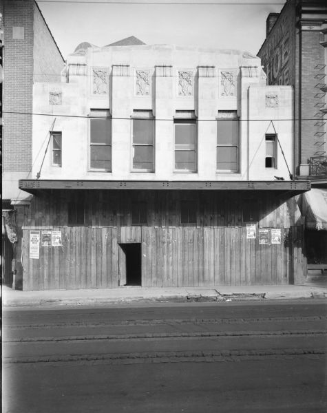 Exterior view from street towards the front of the Orpheum Theater under construction, at 216 State Street. Posters on the temporary wood facade are for the Parkway Theatre advertising W.C. Fields in "So's Your Old Man," and the Madison Theatre advertising Tom Mix in "Hard Boiled." A sign painted on the brick wall of the left side of the building next door on the right reads, in part: "Family [S]hoe [St]ore."