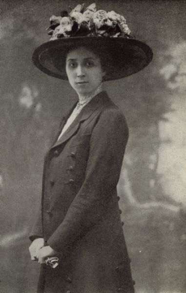 Three-quarter length studio portrait in front of a painted backdrop of Laura M. Cornelius. She is wearing a long coat and a hat with flowers.