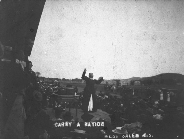 View of (Carry) Carrie Nation speaking from a stage surrounded by a crowd at a county fair.