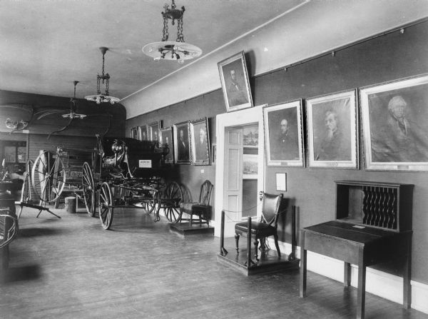 Interior view of a room displaying a carriage, spinning wheel, and two chairs. Paintings are on display, hanging from molding along the walls.
