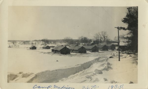 View of several cabins in winter. A path has been cut through snow to the camp. This is Camp Madison, the Civilian Conservation Corps (CCC) camp located at the Arboretum of the University of Wisconsin. The camp was designed by A.E. Galistel.