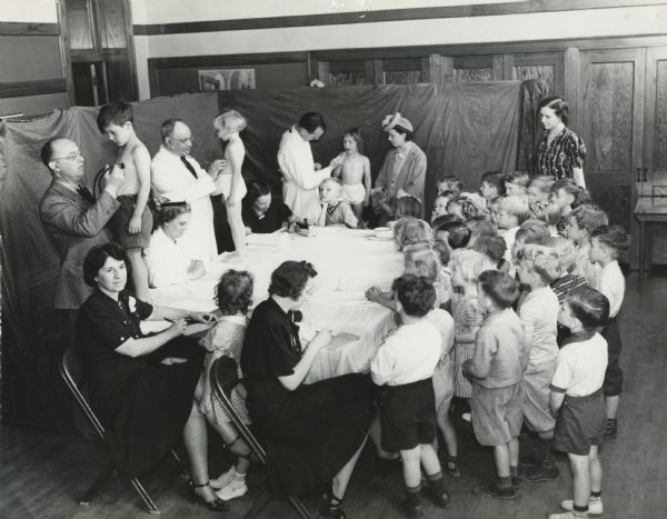 Two men are checking children with stethoscopes, another man is looking into a girl's mouth, and two women are writing in notepads. Other women and many children are standing nearby. Caption reads: "This scene is re-enacted daily in the Town of Lake public health clinic made possible by the WPA [Works Progress Administration] County Public Health Project. A complete history including questions on vaccinations, diseases, family history, etc., is taken of every child who is brought to the clinic."