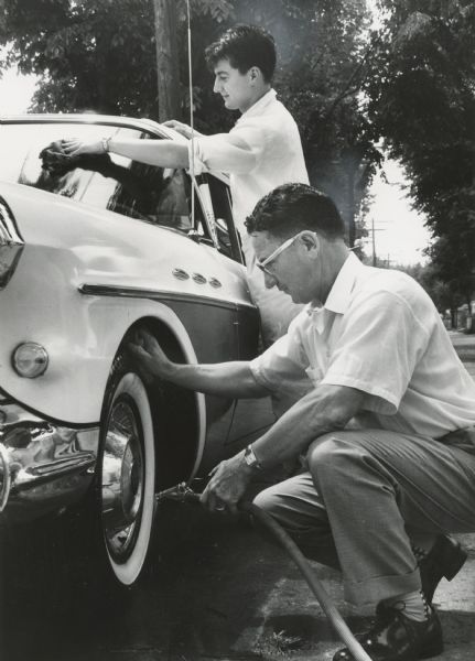A man is spraying his car's front tire rim with a hose, as a younger man is washing the windshield. Caption reads: "<b>Washing his car,</b> Mr. Harger enlisted the aid of his future son-in-law, Andre Winandy. The car had to be spotless for the wedding day activities."