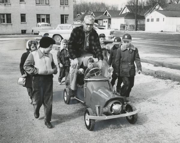 A man is sitting on the rear of a toy car, which is being driven by a girl. Several children are walking alongside the car. Caption reads: "<b>The most popular man</b> in the neighborhood with the children is Earl G. Scherbert of 4105 N. 26th St., and why not? Scherbert, a garage man, built a little automobile powered by a gasoline engine. He gives rides to youngsters on a parking lot evenings and Saturdays when he has time. It is not unusual for him to ride children around for a couple of hours. Scherbert used old auto fenders, channel iron, wheelbarrow wheels and bicycle parts to make the car. For safety's sake, Scherbert rides along on the rear fenders of the car while the youngsters steer."