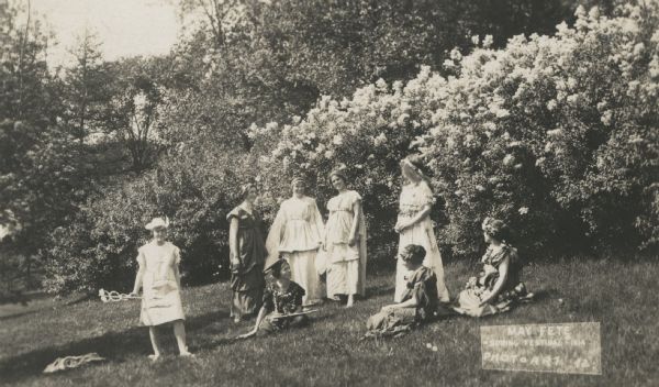 Eight women in costumes are posing in a field, in front of flowering bushes. One is holding a caduceus. Caption identifies this as: "May Fete, Spring Festival 1914."