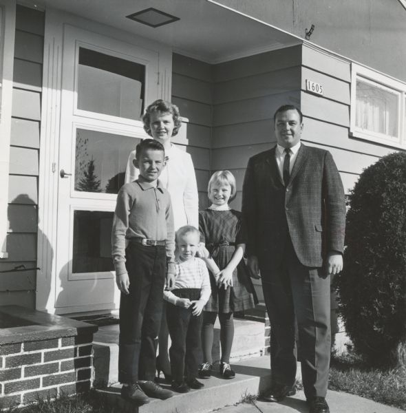 A family with a man, a woman, and three children, are posing in front of a house. Caption reads: "Born on Friday the 13th 30 years ago, Mrs. James [Delores] Lewinski, Marshfield, has abandoned earlier plans to have 13 children. The couple is more than happy to stop with three . . . James, 9; Nancy Jean, 7; and John Joseph, 2. Mrs. Lewinski was the 13th child in her family.