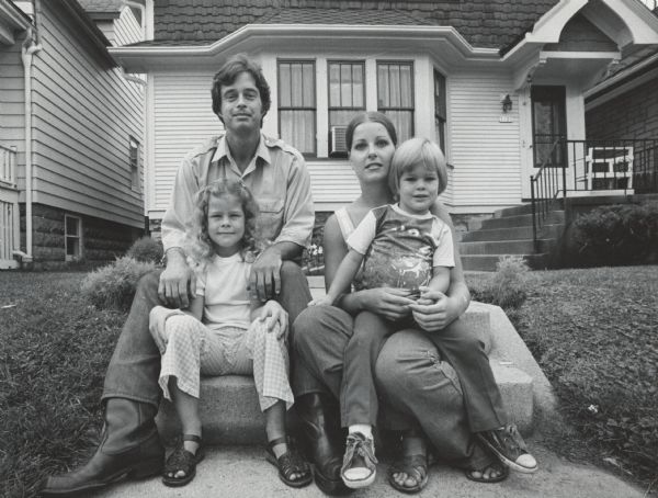 A man, a woman, and two children are sitting together on the front walk steps in front of a house. Caption reads: "Dennis Kubicki, his wife, Sandy, and their children, Deanna, 6, and Dennis Jr., 4, sat outside their home on Milwaukee's South Side. The family has been affected by the 11 week strike by 525 shopworkers against the George J. Meyer Manufacturing Co. in Cudahy. Kubicki is a production electrician."