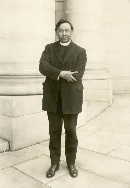 Full-length portrait of Father Philip Gordon standing near a column with his arms crossed.