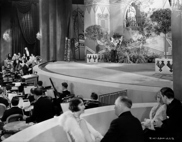 A man with a guitar is standing on the stage in a scene from the 1929 film "Alibi." He is looking up at a woman standing on a balcony above him. The orchestra is in a pit in front of the stage, and two couples are sitting in a box seat near the stage.
