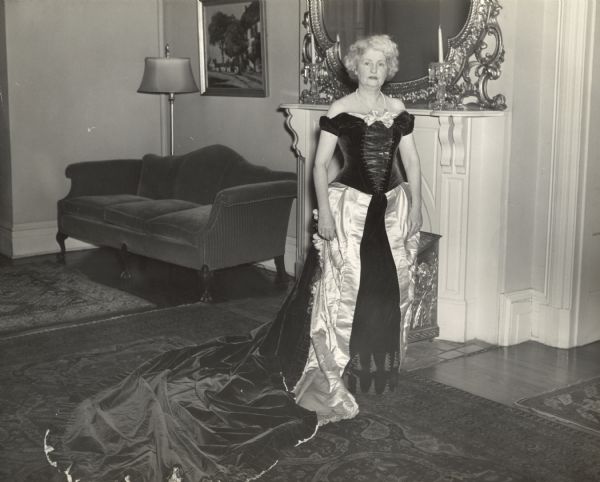 A woman is standing and posing in a formal dress. Caption reads: "Mrs. L[ucius] [Frances] Fairchild's Court Dress worn by Mrs. Walter [Madge] Goodland, Gov. wife. Dark purple velvet and lavender satin worn by Mrs. Fairchild at the Courts in England and Spain. Made by Worth of Paris in 1880. Violet satin shoes, violet kid gloves, violet silk hose and a white silk veil served as accessories."