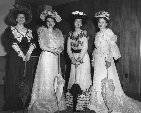 Four women are posing in costume. Caption reads: "The 'Minorettes' Quartet who sang for the 50th anniversary banquet of the Madison Art Association Memorial Union (Great Hall), 5/1/51:  The museum staff helped dress girls in the museum costumes: From left to right: Mrs. J.W. Coyne (Rosemary), 104 S. Brooks, Madison is wearing dress 46.420 - black silk with red trim. 46.467 - large red hat, 1895. Also red parasol. Miss Arlene Radl: 48.1016 - garden party dress, 1908. 48.1140 - grey net hat with flower trim. 46.432 - light or dove blue ostrich feather boa. - Pink parasol with floral border. Mrs. Dorothy Bleicher: 48.1173 - 2 piece white wool and lace dress, sequin trim, c. 1900. 46.473 - lace hat trimmed with pink and red roses. - ecru lace parasol. (Miss Radl has someone take picture [sic] with her camera. She works for Bill Wollin's photo studio.)"