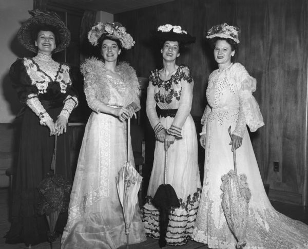 Four women are posing in costume. Caption reads: "The 'Minorettes' Quartet who sang for the 50th anniversary banquet of the Madison Art Association Memorial Union (Great Hall), 5/1/51: 
The museum staff helped dress girls in the museum costumes:
From left to right:
Mrs. J.W. Coyne (Rosemary), 104 S. Brooks, Madison is wearing dress 46.420 - black silk with red trim. 46.467 - large red hat, 1895. Also red parasol.
Miss Arlene Radl: 48.1016 - garden party dress, 1908. 48.1140 - grey net hat with flower trim. 46.432 - light or dove blue ostrich feather boa. - Pink parasol with floral border.
Mrs. Dorothy Bleicher: 48.1173 - 2 piece white wool and lace dress, sequin trim, c. 1900. 46.473 - lace hat trimmed with pink and red roses. - ecru lace parasol.
(Miss Radl has someone take picture [sic] with her camera. She works for Bill Wollin's photo studio.)"