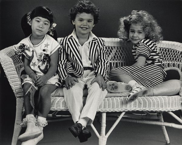 Three children are posing together on a wicker bench. Caption reads: "The nautical look is an important spring theme in children's wear, and it could mean the use of stripes in navy and white accented with red, or an outfit in black and white combined with touches of bright yellow. They also could include (from left) this knit top and short set, about $43, from Brigadoon; a striped jacket, $30, classic white pants, $15, and a white shirt, $10, from The Children's Place; or the tried-and-true look of a striped knit dress, $34, from Louise Godell. Other fun looks include cropped tops and clam-digger pants in bright prints, says B.J. Rafenstein, owner of Louise Goodell, 314 E. Silver Spring Dr., Whitefish Bay."