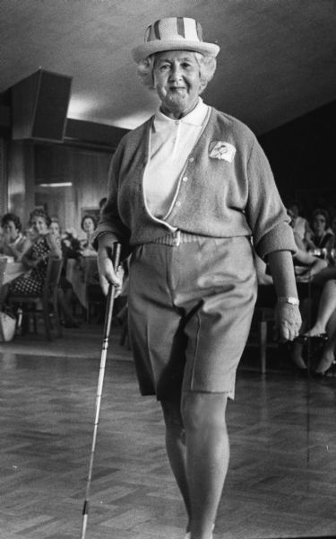 A woman is posing in an outfit, holding a golf club. A large group of women are sitting in chairs at tablesin the background. Caption reads: "<b>Today's golfer</b> would be stylish in shorts, sweater and straw hat similar to those modeled by Mrs. Jay [Doris] Minow, 1202 E. Kenmore pl., Shorewood."