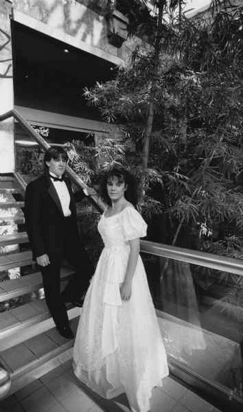 A young man in a tuxedo and a young woman in a dress are posing on a staircase, with a bamboo plant behind them. Caption reads: "Courtney Thomey and Jenni Pemberton model prom outfits that will be auctioned Saturday evening at Mayfair Mall Atrium."