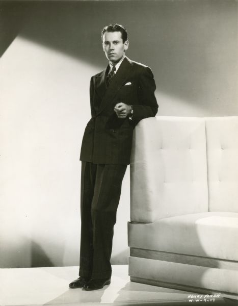 Full-length publicity portrait of Henry Fonda. He is standing and leaning on a high-backed white sofa. He is wearing a dark suit and has a blank look on his face.