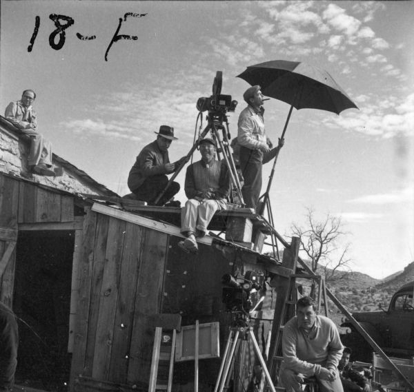 Director Herb Biberman and three other men are sitting and standing on a platform built on the roof of a shack during the filming of "Salt of the Earth." Biberman is sitting underneath the camera. A man is sitting next to another camera on the ground below them. "18-F" is written in the top left corner of the photograph.