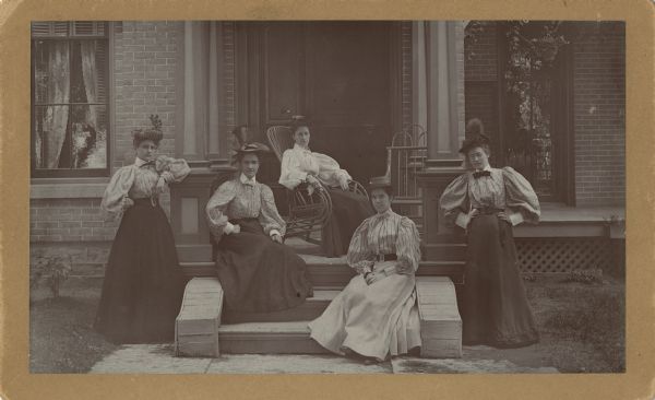 Five women are posing in front of a house on a stoop and porch. They are all wearing hats, skirts, and blouses, and one woman is sitting in a rocking chair. Caption reads: "Parkinson home on Ludington St - Columbus, Wisconsin."