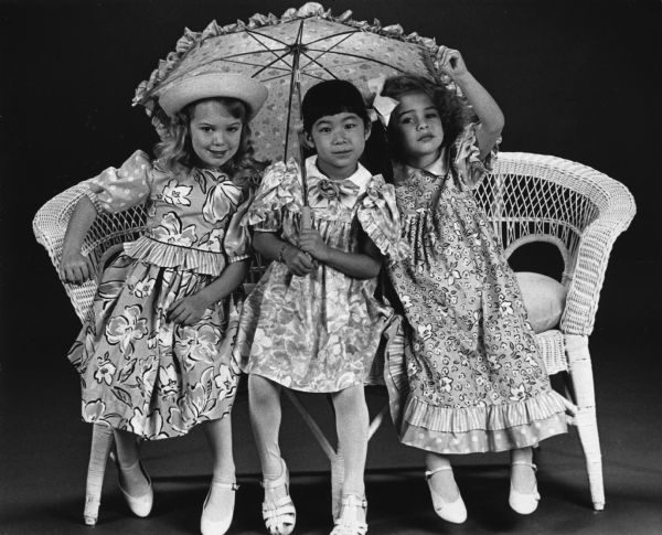 Three girls are sitting on a wicker chair, and wearing floral print dresses. The middle girl is holding a parasol over the three of them. Caption reads: "Floral print dresses in pastel colors are among the newest dressy trends for spring for girls sizes 4 to 6X, according to Diane Long, manager of The Children's Place at Northridge Shopping Center. Shown (from left) are a lavender/pink dress, about $103 from Brigadoon; a lavender/blue print dress, $45 from Marshall Field's; and a blue print dress, $58 from Louise Gooddell. Floral prints also are popping up in sportswear, said Terri Luljak, co-owner of Brigadoon, 11033 N. Port Washington Rd., Mequon."