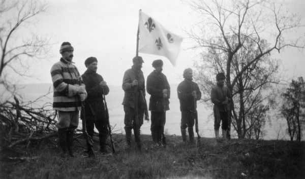 Six actors are standing outdoors. One of them men is holding a flag a fleur-de-lis design in a reenactment of a ceremony in which Nicolas Perrot claimed the territory of the upper Mississippi River for France at Fort St. Antoine.