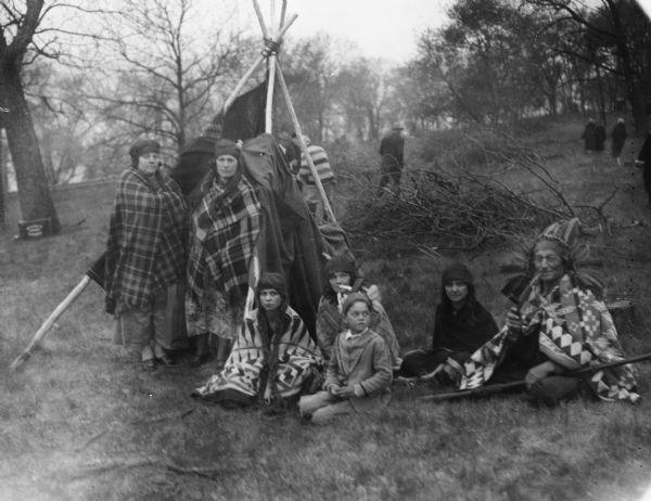Actors (who are possibly Native American) posing outdoors near a lodge. They are participants in a reenactment of a ceremony in which Nicolas Perrot claimed the territory of the upper Mississippi River for France at Fort St. Antoine.  