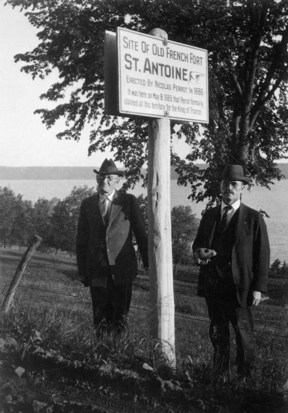 Two men are standing by a sign, near a fence, marking the former location of Fort Antoine.