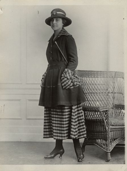 A woman is standing and posing near a wicker chair. She is wearing a hat, coat, skirt, and has a cane tucked under her arm. Caption reads: "Smart hiking suit in dark green velour in combination of half inch check in green and grey."