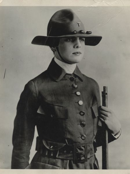 Waist-up portrait of a woman wearing a uniform and holding a rifle. Her helmet has a pin with the caduceus, as was used on the United States Army Medical Department uniform. Caption reads: "Military uniform of khaki which is now being worn by Women all over the country at their training camps and to stimulate recruiting."