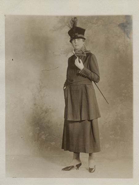 Full-length portrait in front of a painted backdrop of a woman wearing a hat and dress, and holding a cane under her arm. Caption reads: "Straightline suit of navy serge trimmed with much soutache braid and finished with a grey velvet tippet about the neck."