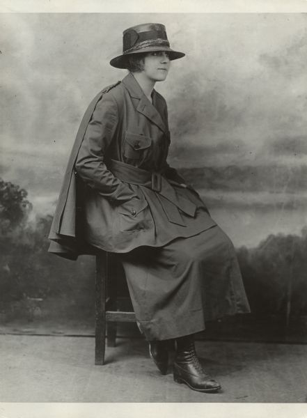 Full-length portrait in front of a painted backdrop of a woman posing sitting on a chair and wearing a hat, coat with cape, and skirt. Caption reads: "Trench togs are all the go now and this short skirt and soldierly coat, to say nothing of the detachable cap [cape?], are of cotton khaki for service."