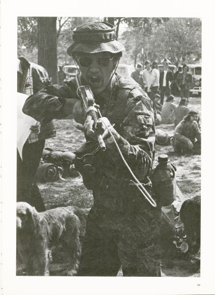 A Vietnam veteran is pointing a gun toward the camera. He is wearing sunglasses, a hat, and camouflage. There is a dog standing beside him on the left. People are sitting and standing in the background.