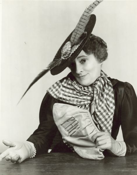 Actress Ruth Draper looking at the camera with a smile. She is wearing a flat hat with two pheasant feathers on it, along with a large checkered scarf and gloves, while holding a purse with the initials "FBG" on it. The photograph is from her appearance at the Empire Theatre in January and February 1947.