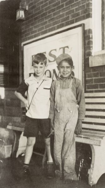 Two boys are standing outdoors. Behind them is a bench along the side of a brick building.