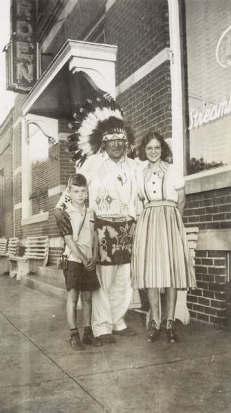 Two young people posing with a Native American man in a feather headdress and beaded clothing on sidewalk in front of a brick building.