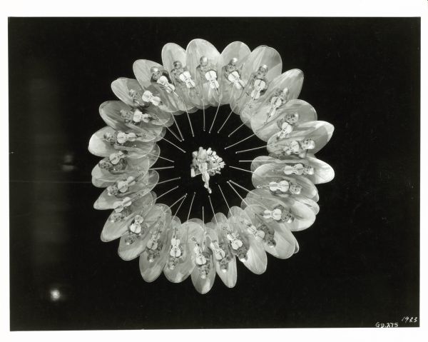 Overhead view of dancers in a circle during the "Shadow Waltz" number in the film "Gold Diggers of 1933." The dancers are each wearing large hoop skirts and are holding violins with the bows pointing to the dancer in the center of the circle.
