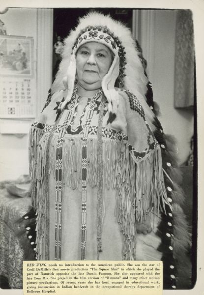 Lillian St. Cyr standing and posing in a traditional Native American beaded buckskin dress and floor length headdress. St. Cyr, also known as Red Wing, was a silent film actress between 1908-1921.