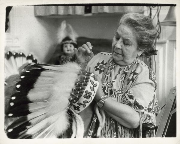 Lillian St. Cyr working on a headdress. The headdress has a beaded headband with feathers attached to it. She is wearing a traditional Native American beaded buckskin dress. St. Cyr, also known as Red Wing, was a silent movie actress between 1908-1921. 