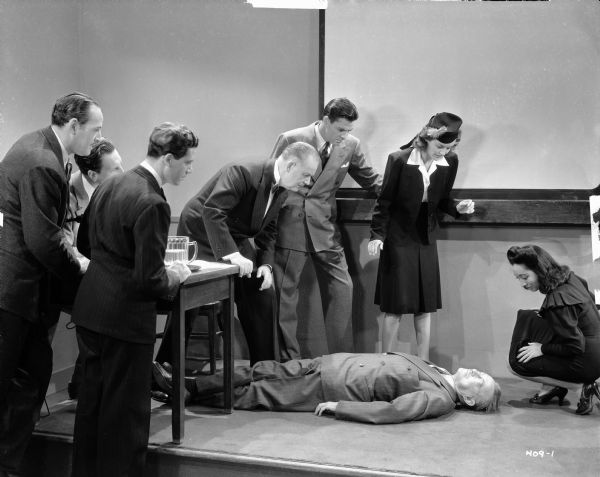 A group of people are standing and looking down at Dr. John Benton who has collapsed and is laying on the floor in a scene from the 1940 film "Phantom of Chinatown."