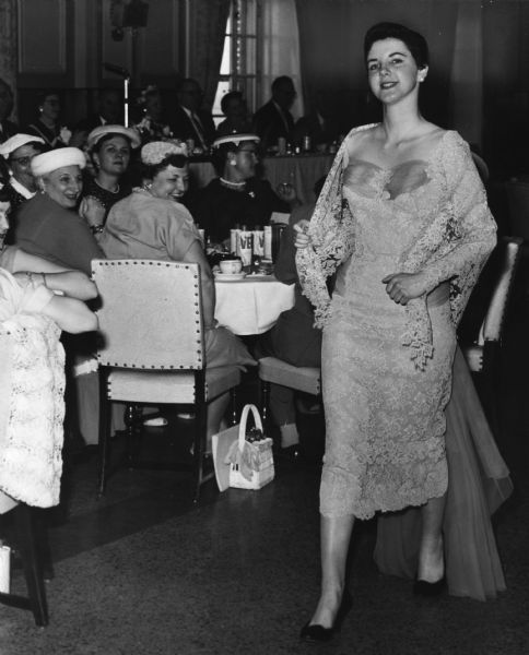 A woman is modeling a dress with a shawl and a train. Behind her women are sitting at tables watching her go by. Caption reads: <b>A dress worn by actress Celeste Holm</b> in the movie "High Society" was modeled by Miss Judith Benning, 2495 S. Delaware Av., at the 25th anniversary luncheon of the Better Films Council of Milwaukee County Monday at the Elks club."